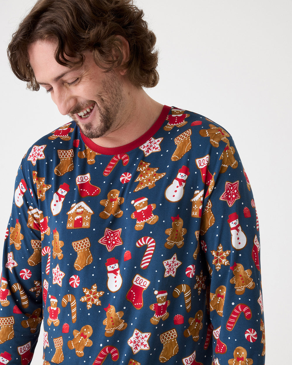 A man wearing a festive Christmas long-sleeve shirt with a blue background and a pattern of gingerbread men, snowmen, candy canes, and stockings, photographed against a plain light gray background.