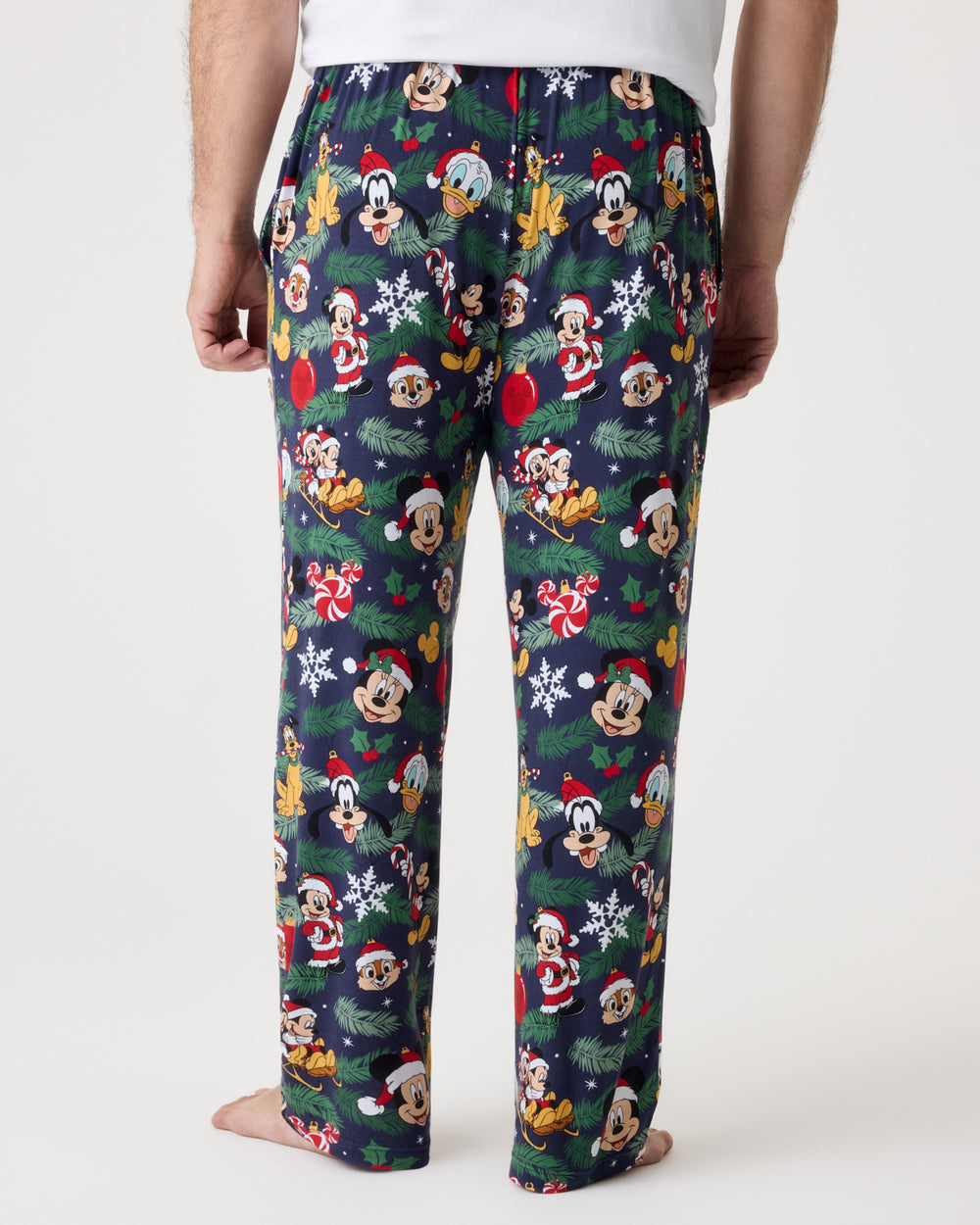 A person shown from the back wearing dark navy Christmas-themed pajama pants covered in a repeating print of Disney characters in Santa outfits, with snowflakes, pine branches, and holiday motifs, against a plain light background.