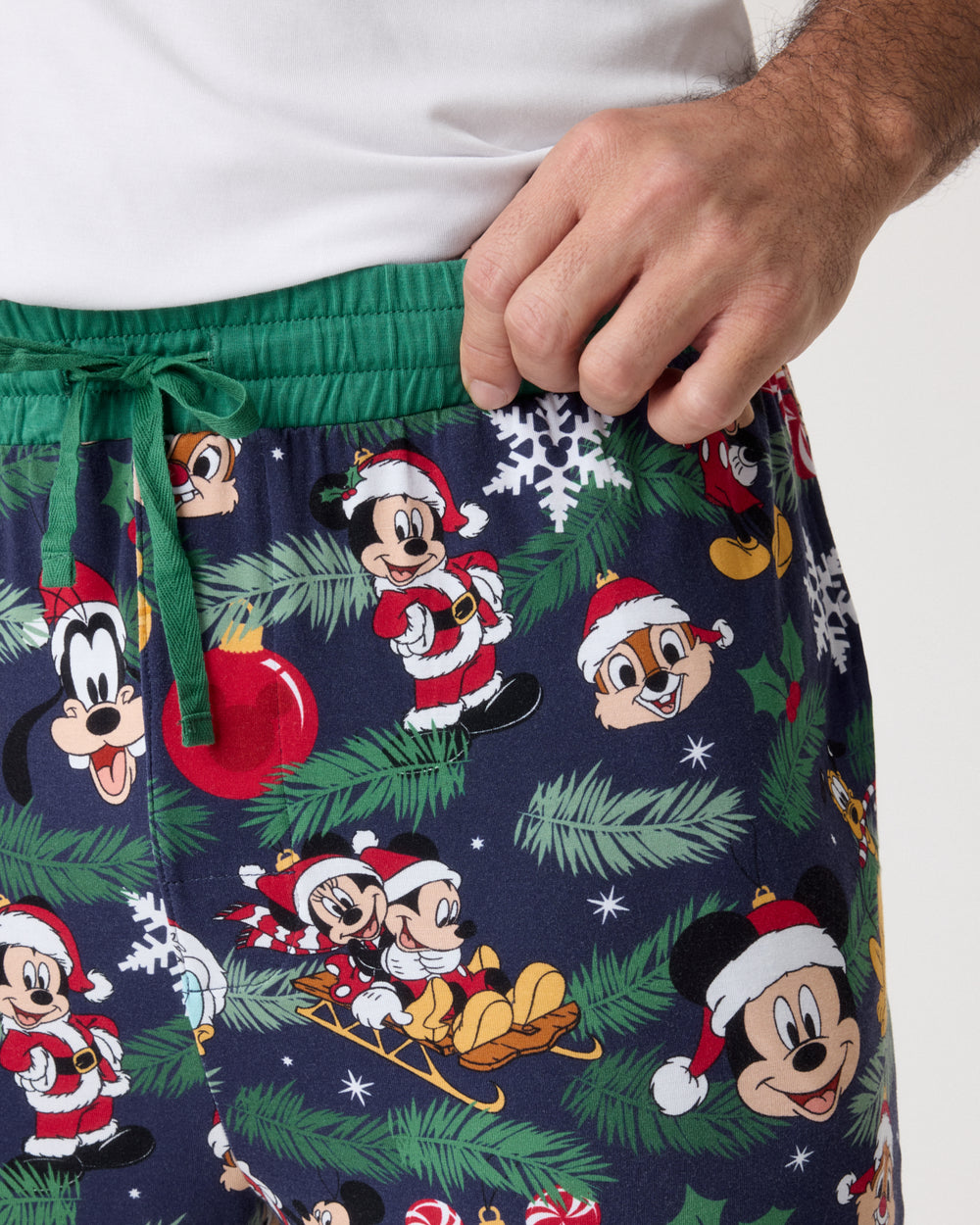 A close-up of a person wearing dark navy Christmas-themed shorts with a green drawstring waistband, featuring a festive print of Mickey Mouse, Goofy, and Chip 'N' Dale in Santa outfits with pine branches, snowflakes, and ornaments. The person is also wearing a white t-shirt.