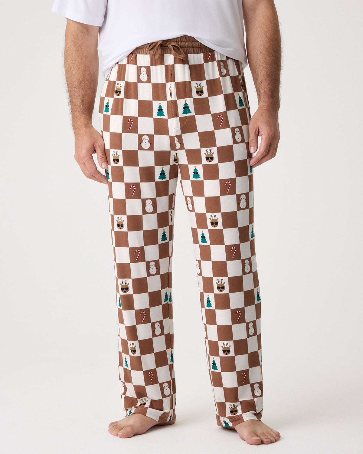 A man wearing brown and white checkered pajama pants with small holiday-themed icons printed on them, paired with a white t-shirt, shown against a plain light background.