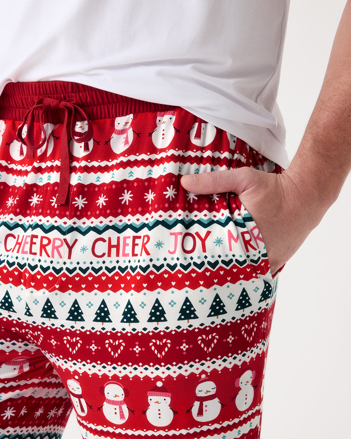 A close-up of a person wearing red and white holiday knit pants with snowman, tree, and chevron patterns, plus the text 'CHEER JOY' across the front.