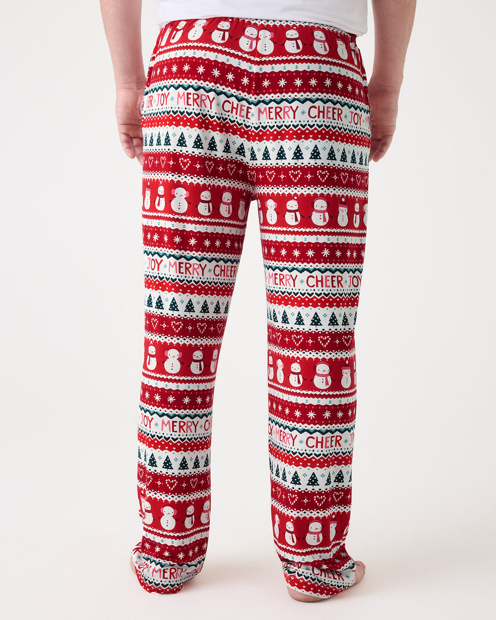 A person shown from the back wearing red holiday pants with a festive pattern of snowmen, Christmas trees, hearts, and the words 'MERRY CHEER' on a light gray studio background.