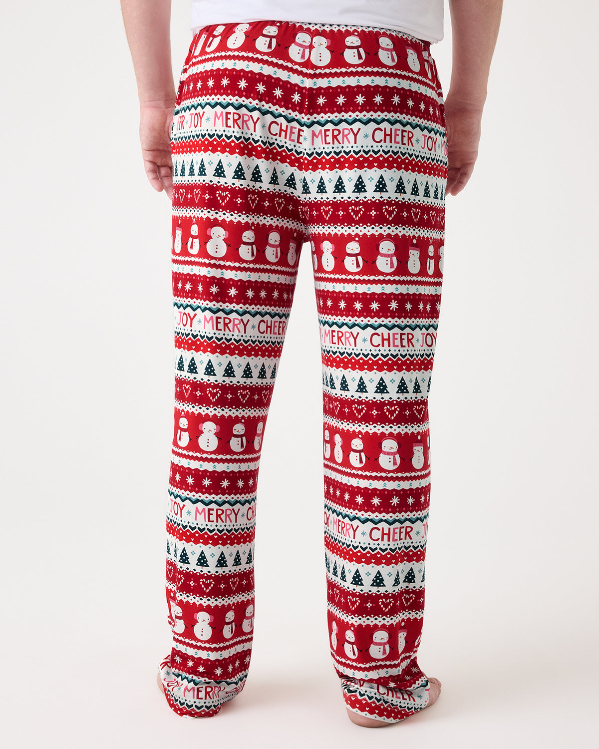 A person shown from the back wearing red holiday pants with a festive pattern of snowmen, Christmas trees, hearts, and the words 'MERRY CHEER' on a light gray studio background.