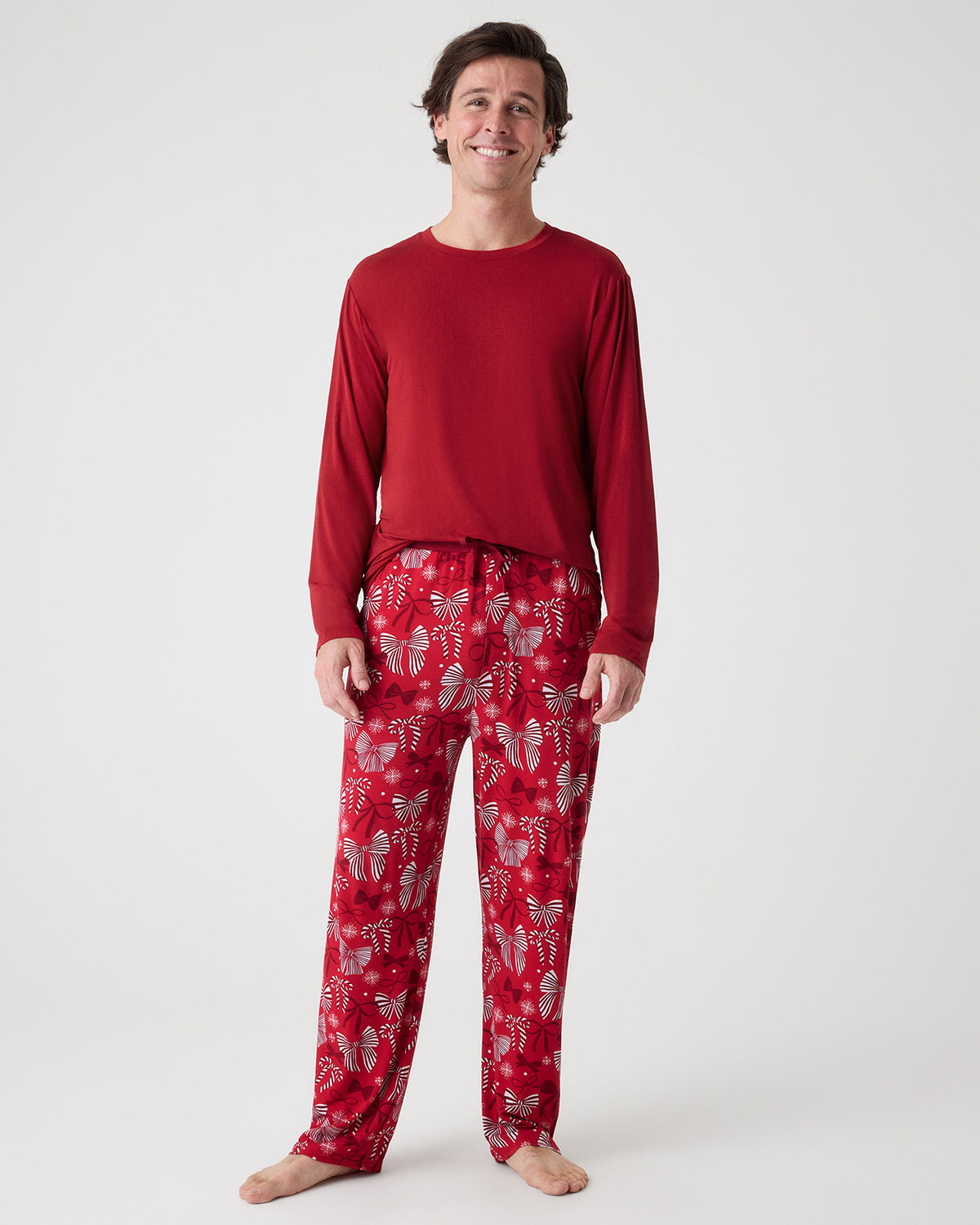 A man modeling a red long-sleeve crewneck shirt and matching red pajama pants with a holiday bow pattern, standing against a light gray studio background.