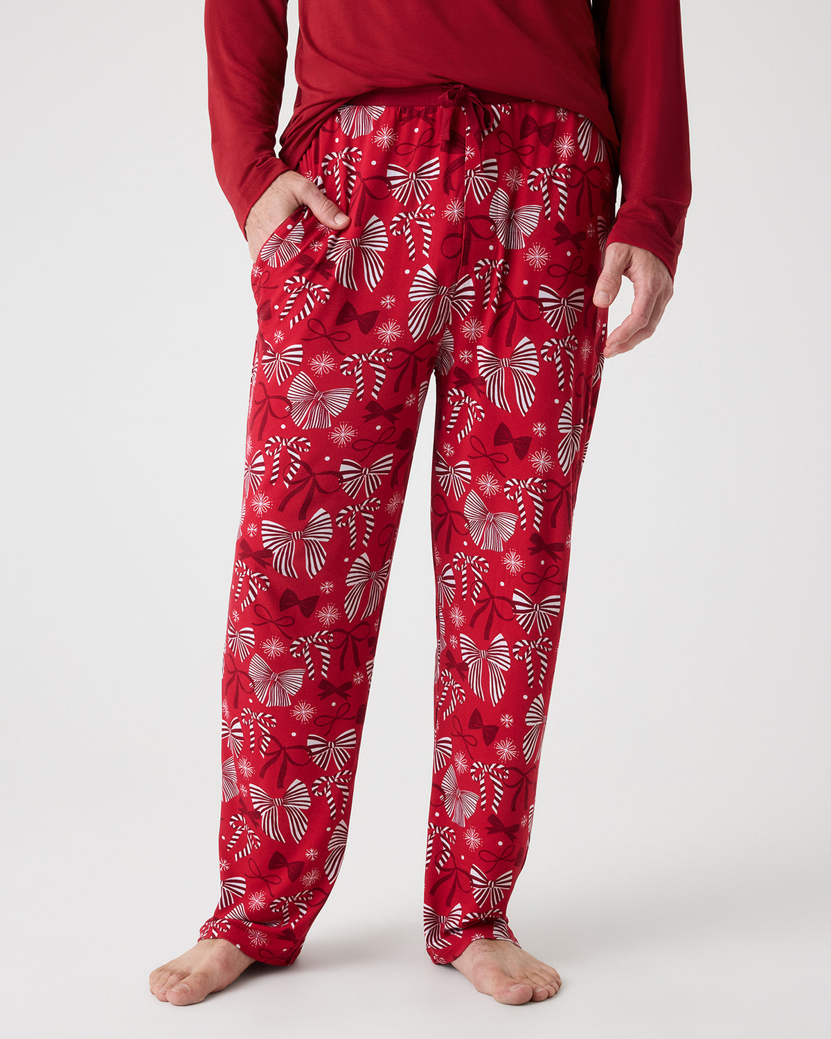A man wearing red pajama pants with a white holiday bow pattern, paired with a dark red long-sleeve top, standing against a light gray studio background.