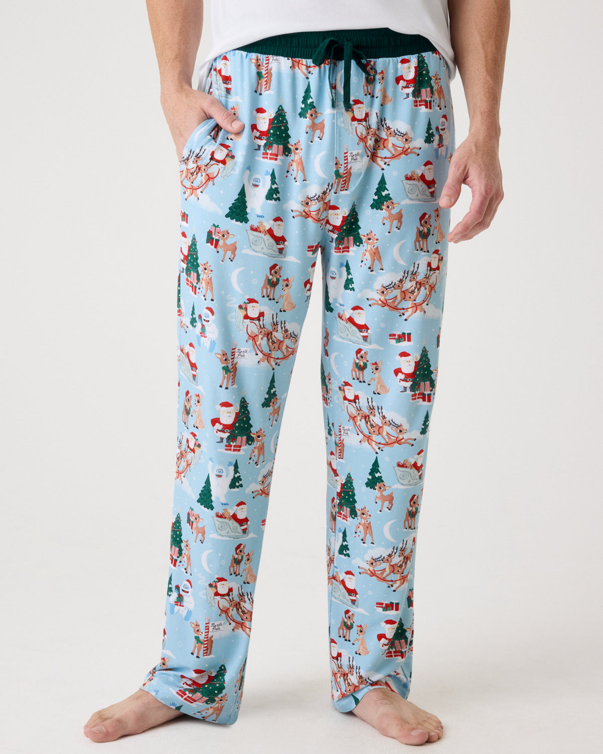 A person wearing light blue holiday-themed lounge pants with an all-over print of Santa Claus, snow globes, Christmas trees, and sleighs, shown from the waist down against a plain light background.