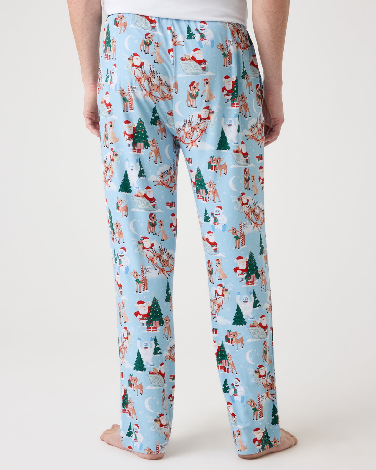 A person shown from the back wearing light blue pajama pants with a festive Christmas print featuring Santa Claus, reindeer, snowmen, and trees, standing barefoot against a plain light background.