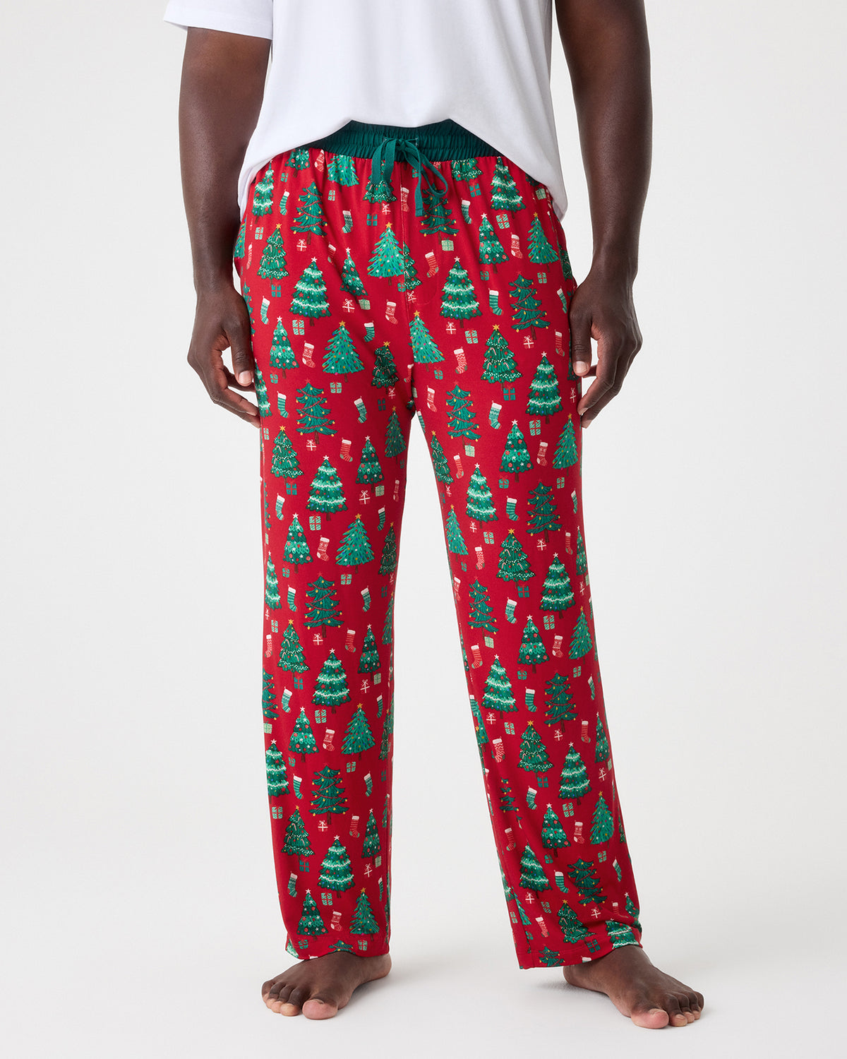 A person wearing red holiday pajama pants with a green Christmas tree pattern, shown from the waist down against a light gray studio background.