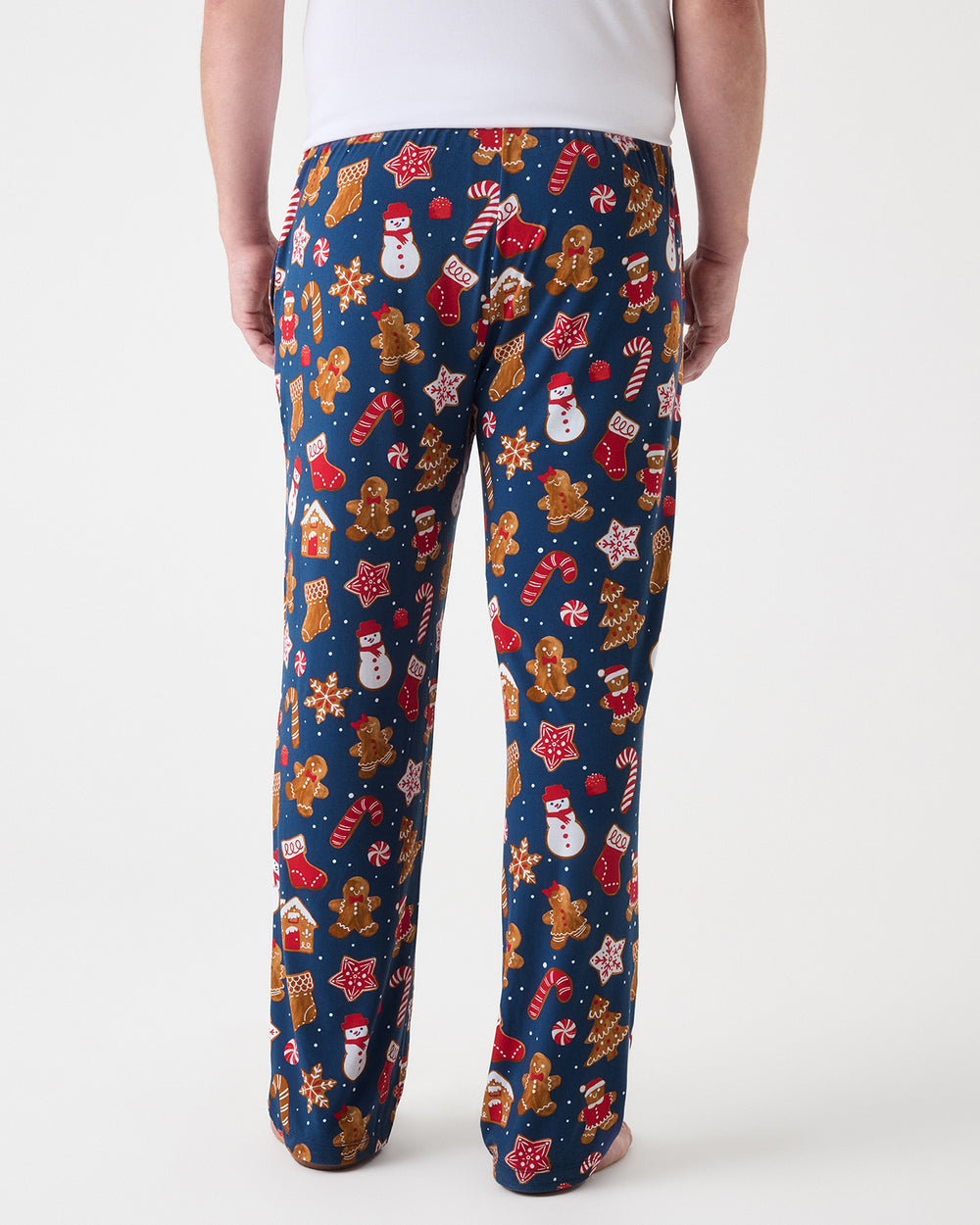 A person shown from the back wearing navy blue holiday print pajama pants with gingerbread men, snowmen, candy canes, and ornaments, against a plain light gray studio background.