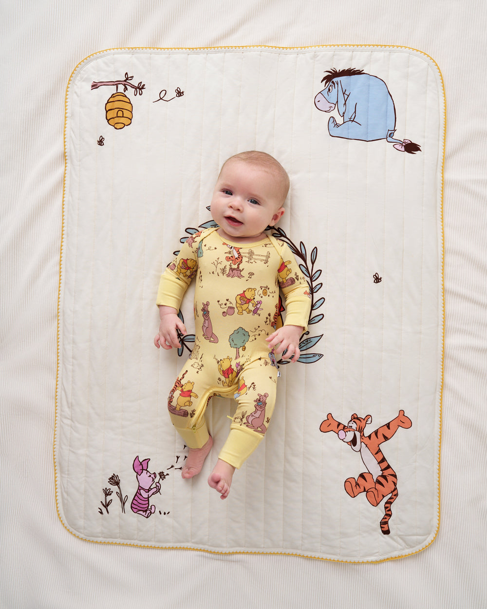 A baby wearing a yellow long-sleeve footless jumpsuit with Winnie the Pooh character prints lies on a white quilted blanket with a yellow border and additional character illustrations.