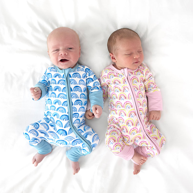 Two infants lying on a bed wearing matching Blue Rainbows and Pastel Rainbows Zippies