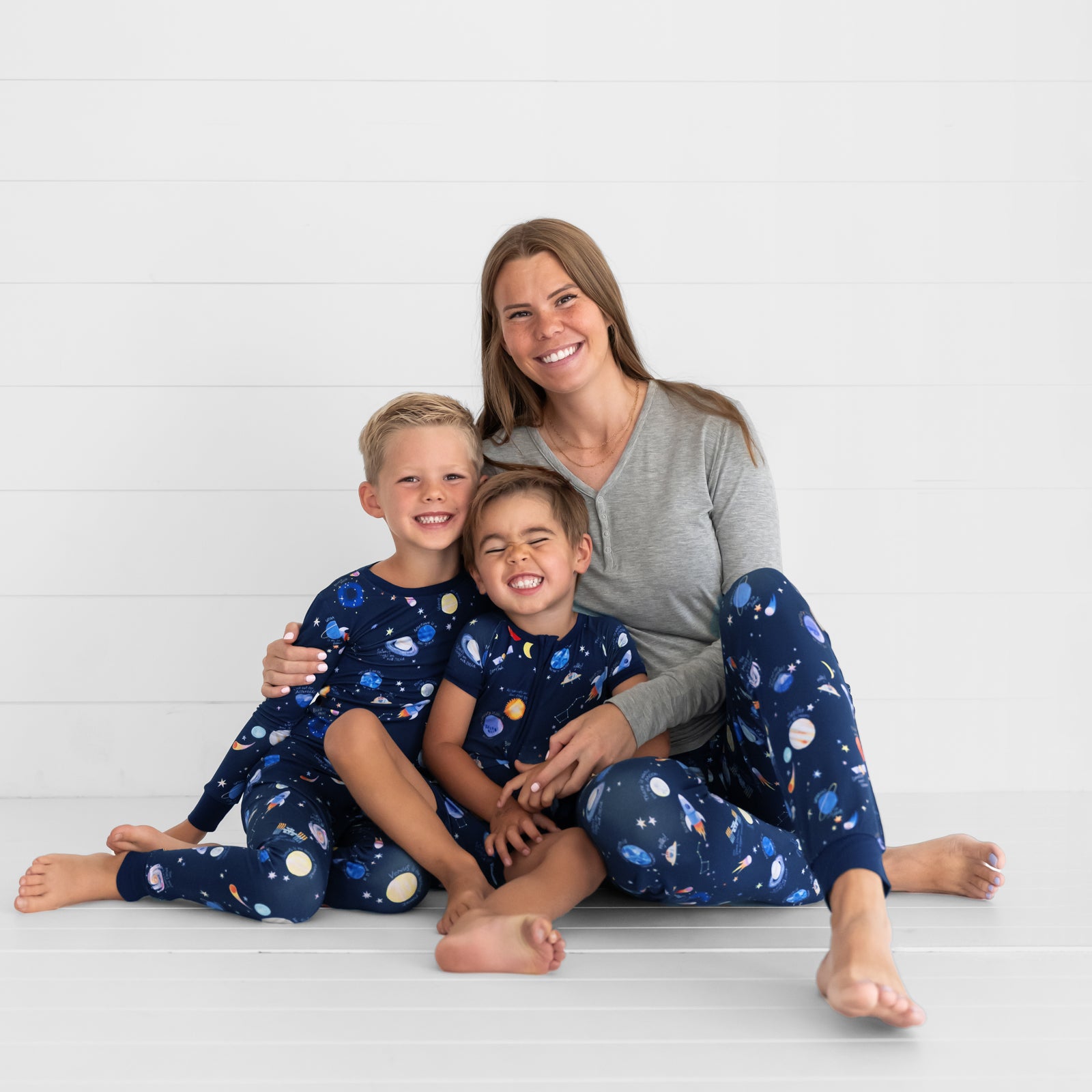 Mother and her two children wearing matching Through My Telescope pjs in women's,  and children's two piece and shorty zippy styles. Mom is matching her pants with a women's Heather Gray pj top