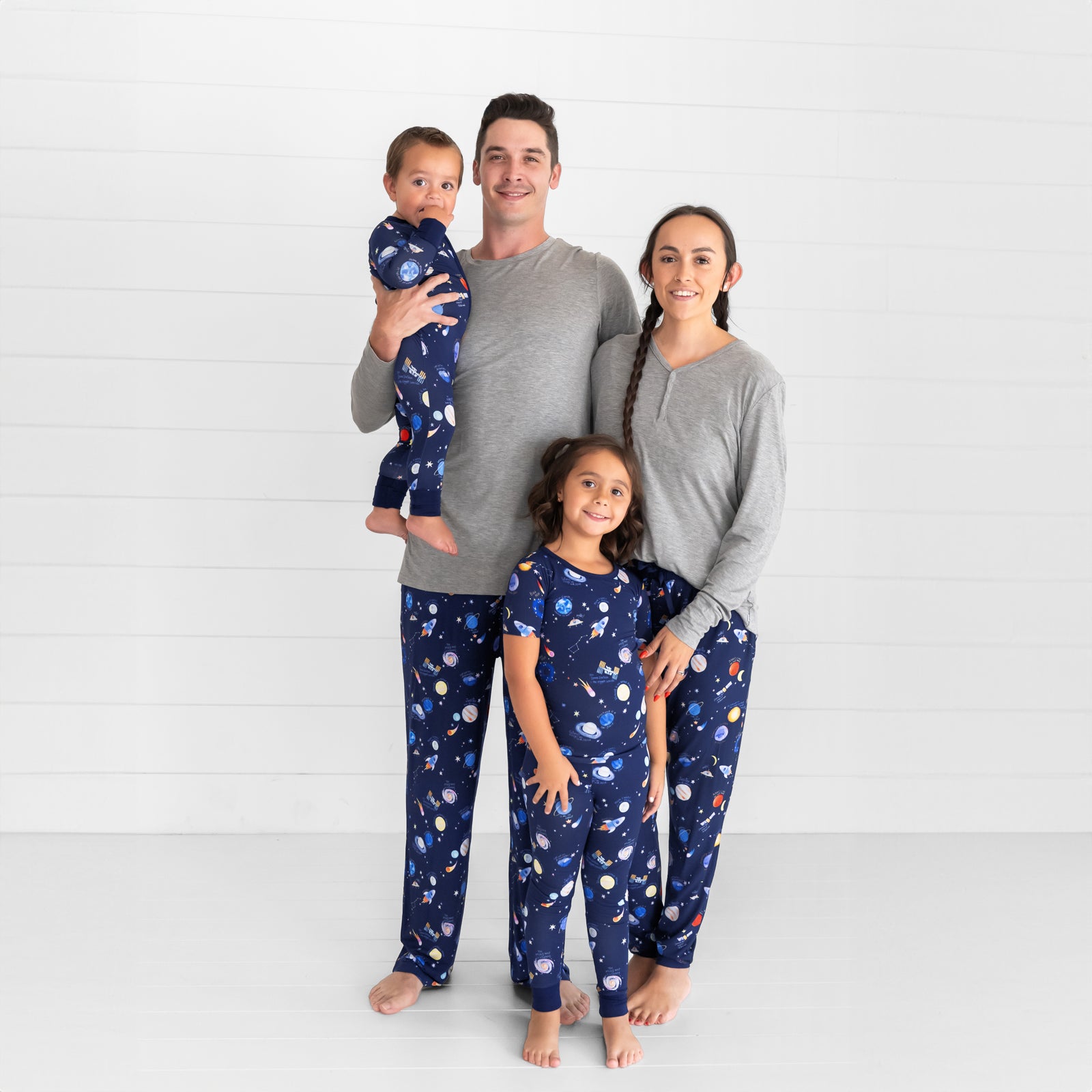 Family of four wearing matching Through My Telescope pjs in men's, women's, and children's two piece and zippy styles. Mom and Dad are pairing their pants with coordinating Heather Gray pj tops in men's and women's styles.