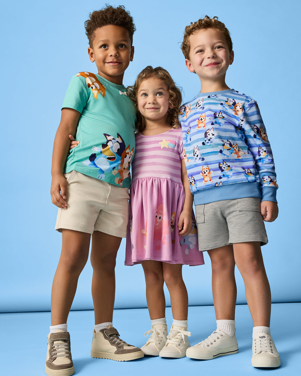 Three children wearing daywear styles from the Bluey Collection on the blue back drop