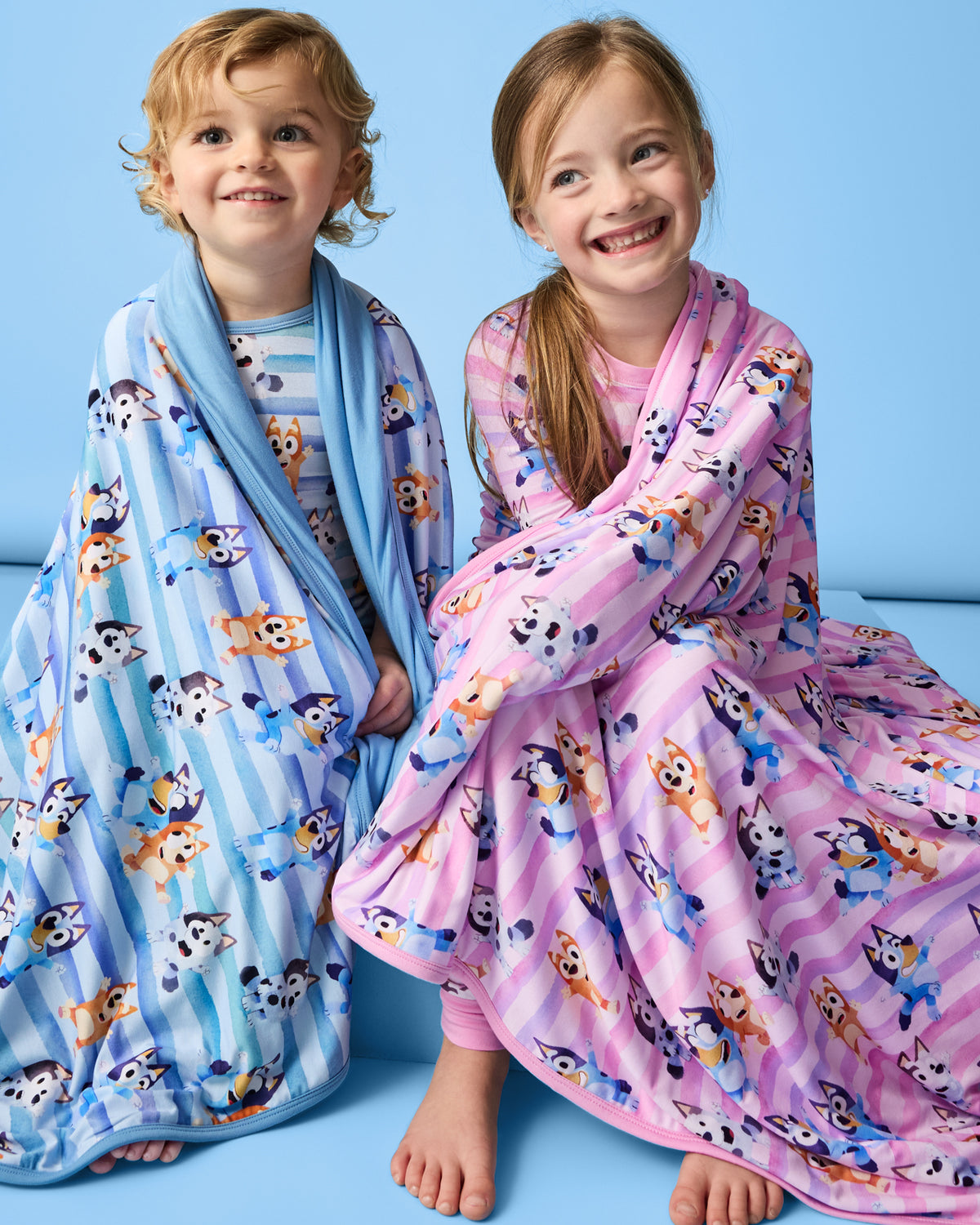 Two children wrapped in Bluey Dance All Day Large Cloud Blankets® on a blue background