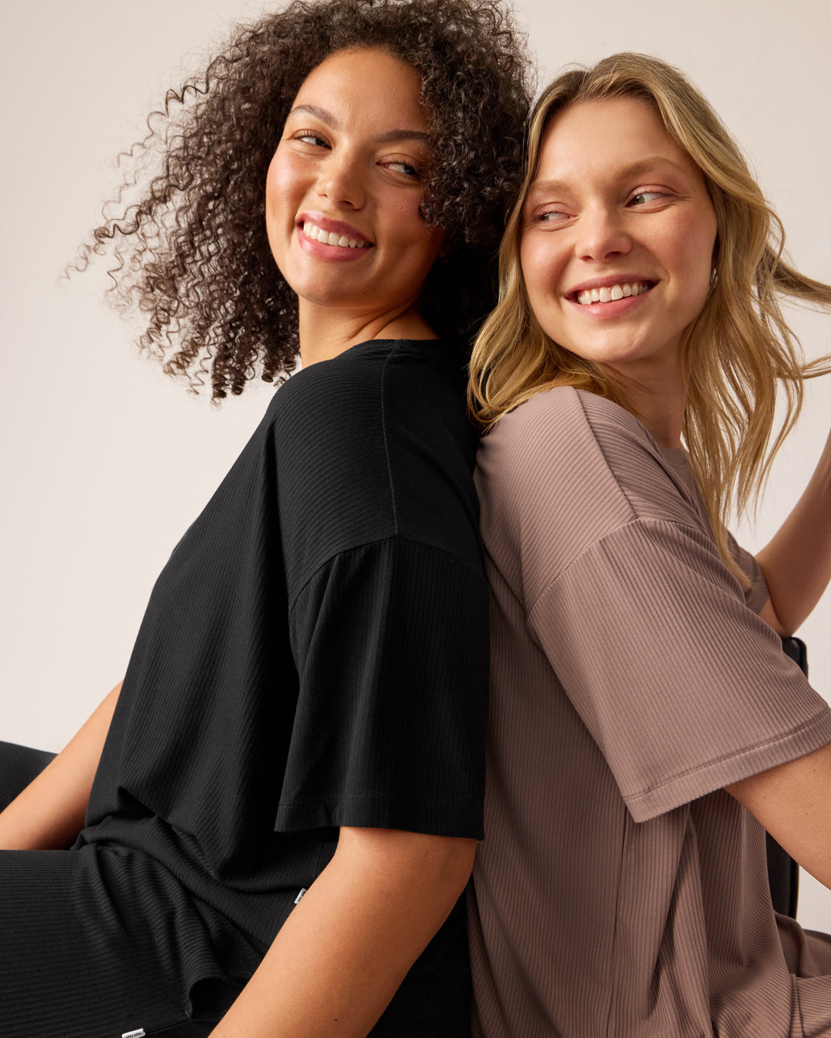 Two woman sitting back to back wearing matching Women's Ribbed Boxy Tees