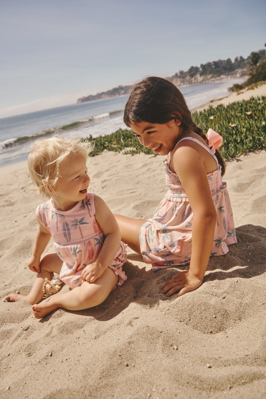 Two children sitting in the sand wearing Pink Palm Dunes summer styles