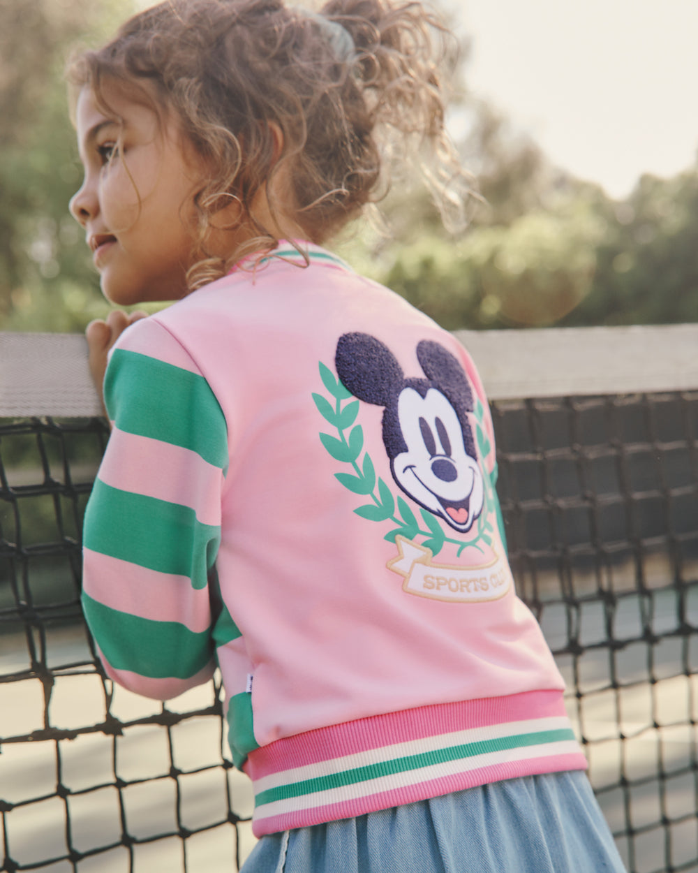 Girl wearing the Disney Mickey Mouse Sports Club Varsity Cardigan leaning on a tennis court net