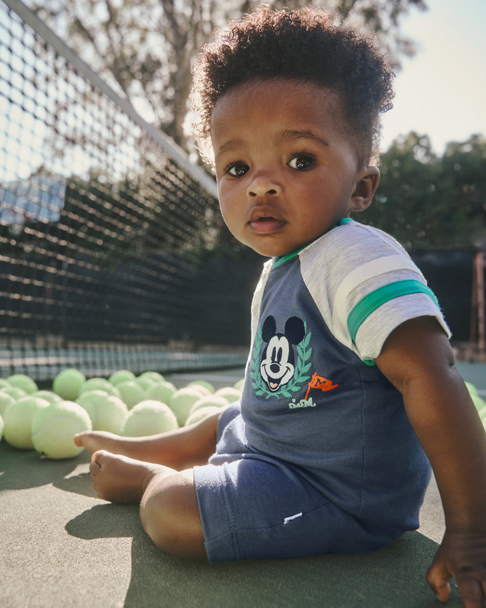 Baby wearing the Disney Mickey Mouse Sports Club Henley Shorty Romper on a tennis court