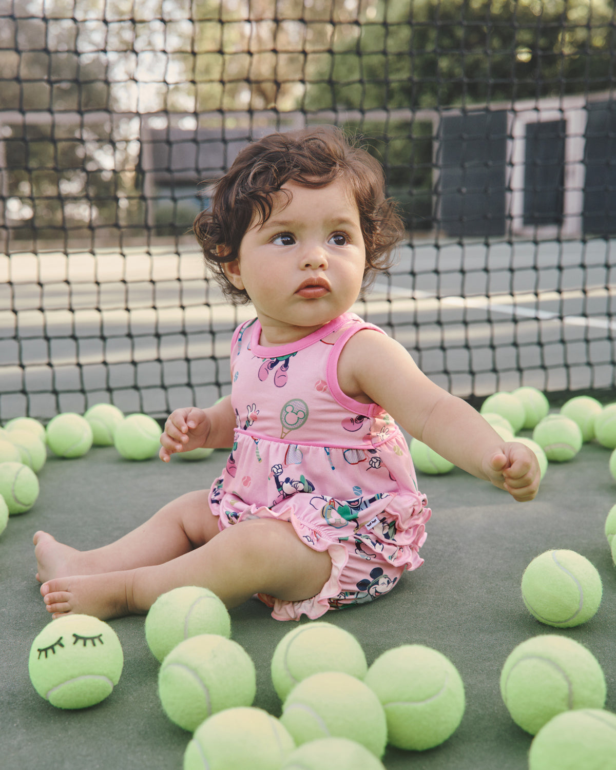 Baby sitting on a tennis court surrounded by tennis balls