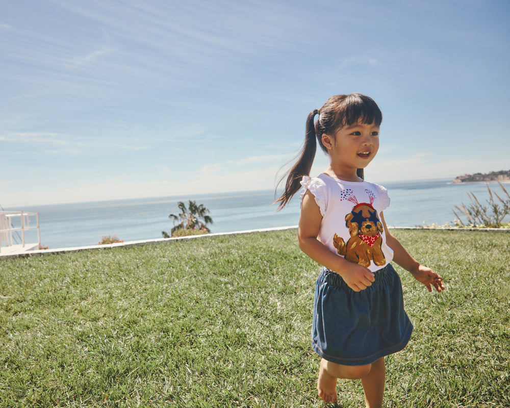 Girl wearing the Star Spangled Pup Flutter Tee and denim skort on a grass lawn
