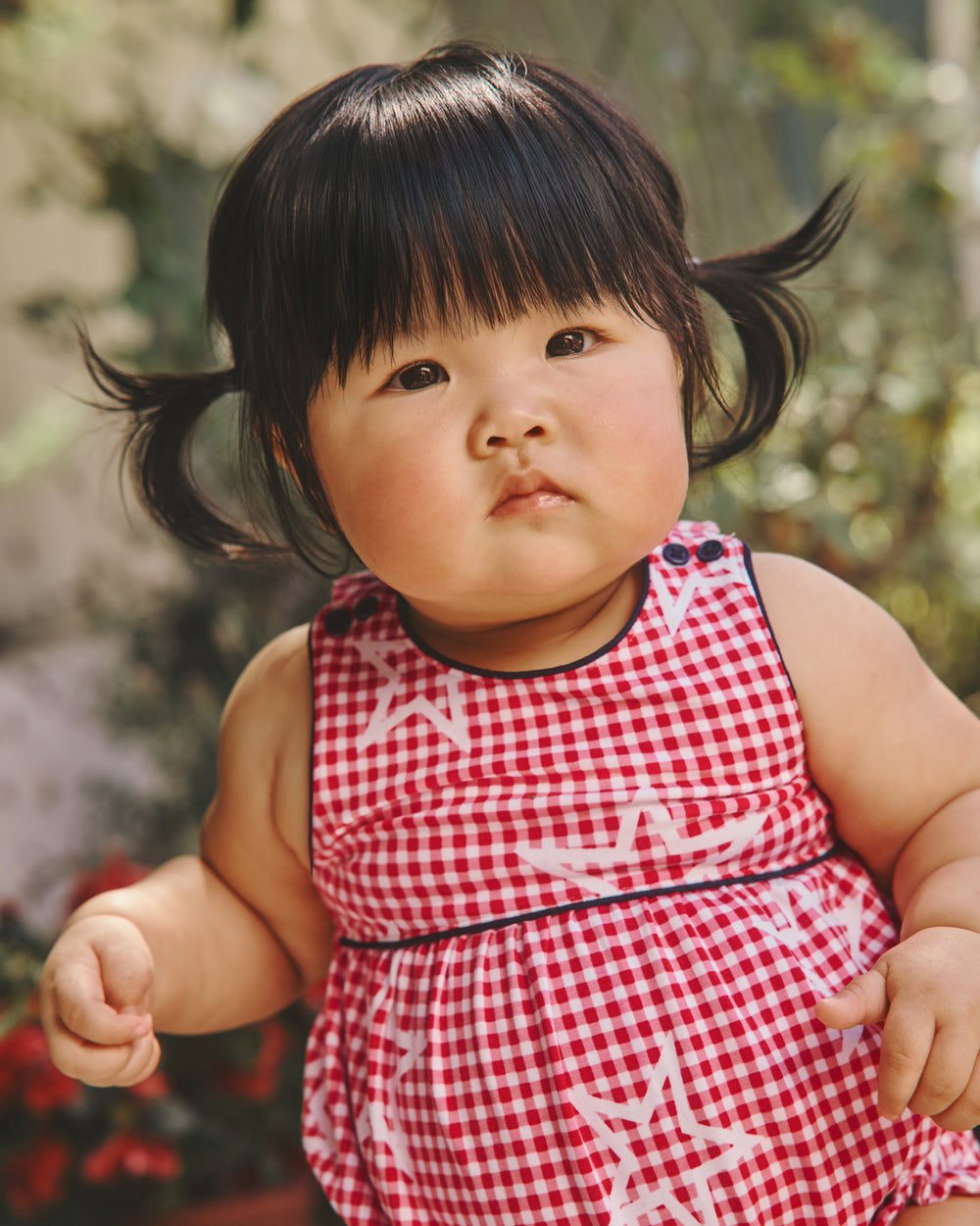 Girl wearing the Celebration Gingham Bow Back Bubble Romper with greenery in the background