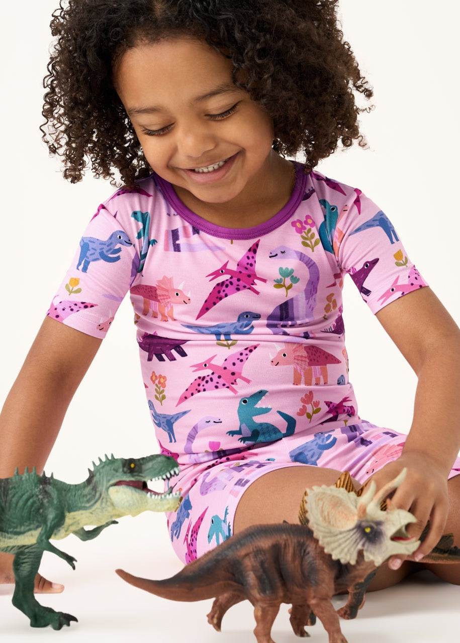 Sitting girl wearing the Pink Roar & Explore Two-Piece Short Sleeve & Shorts Pajama Set playing with dino toys