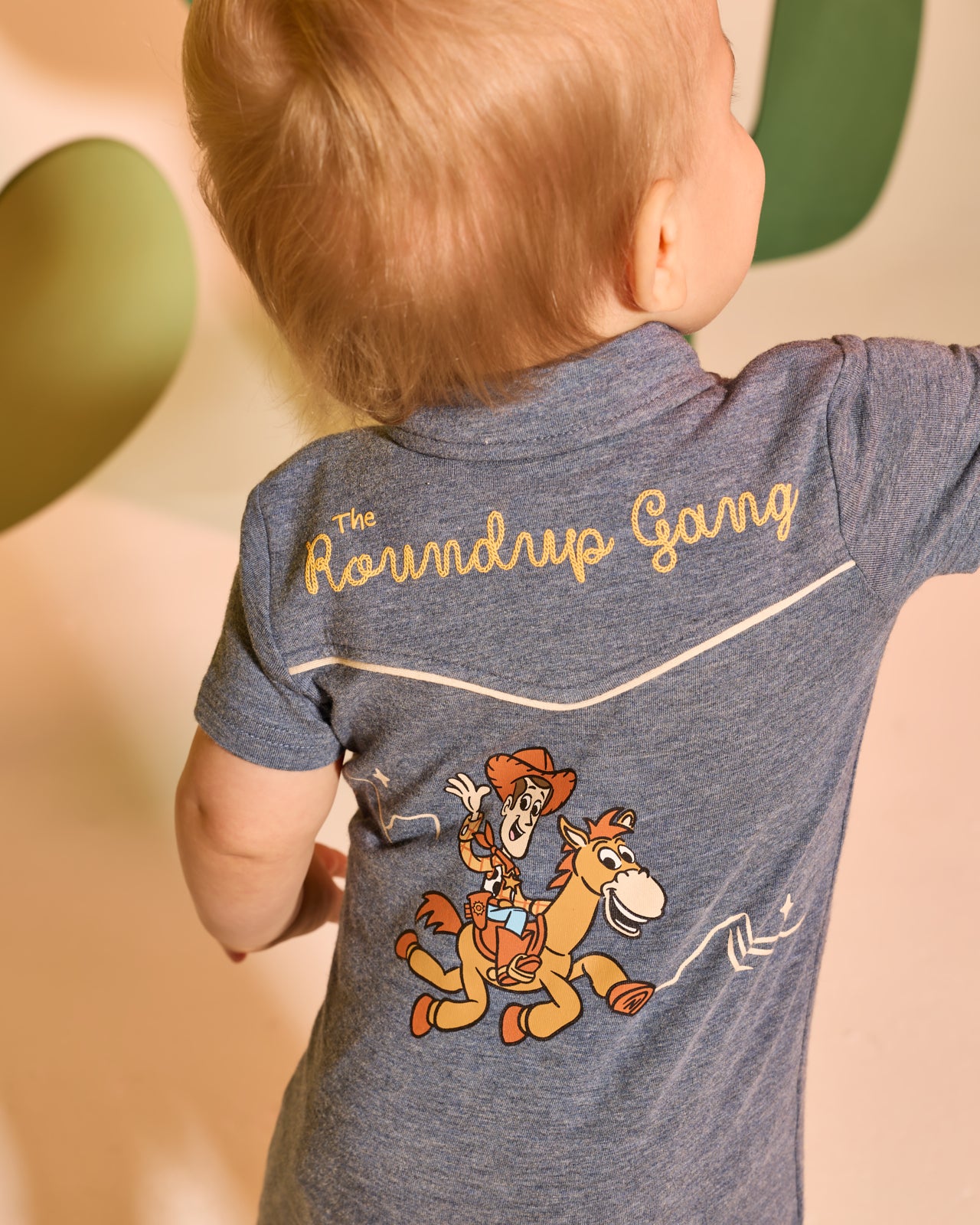 Back view of child wearing the Disney & Pixar The Roundup Gang Polo Shorty Romper on a tan backdrop