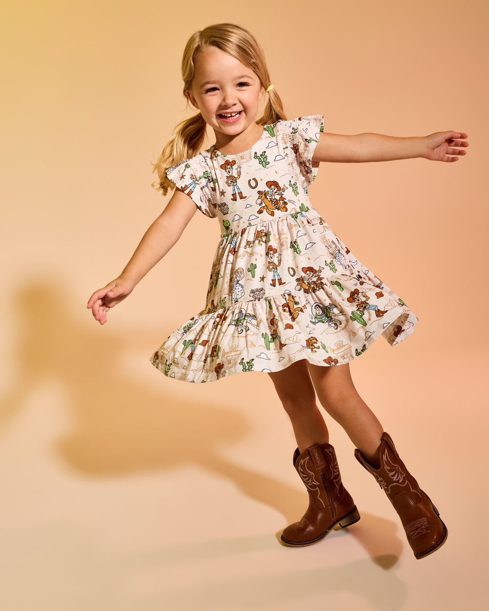 Twirling girl wearing the Disney & Pixar Woody's Wild West Tiered Flutter Dress on a tan backdrop