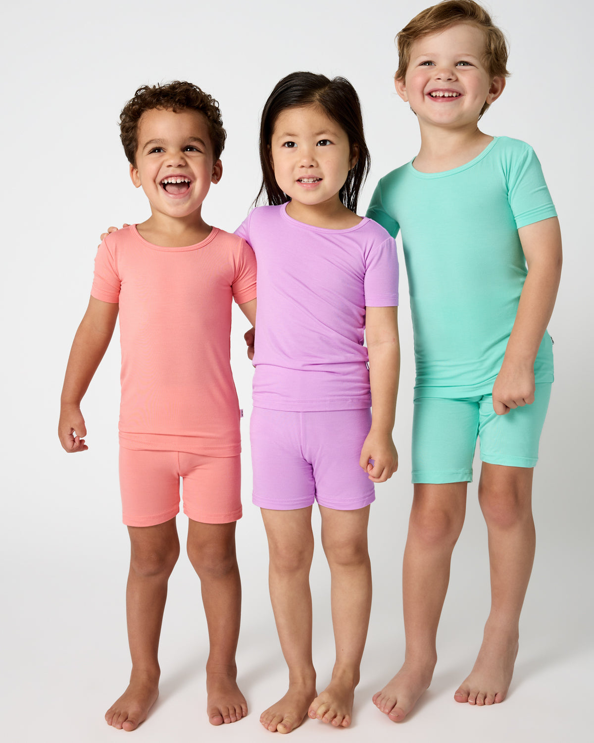 Three kids wearing matching Sweet Papaya, Soft Wisteria and Ocean Mist Two-Piece Short Sleeve & Shorts Pajama Sets
