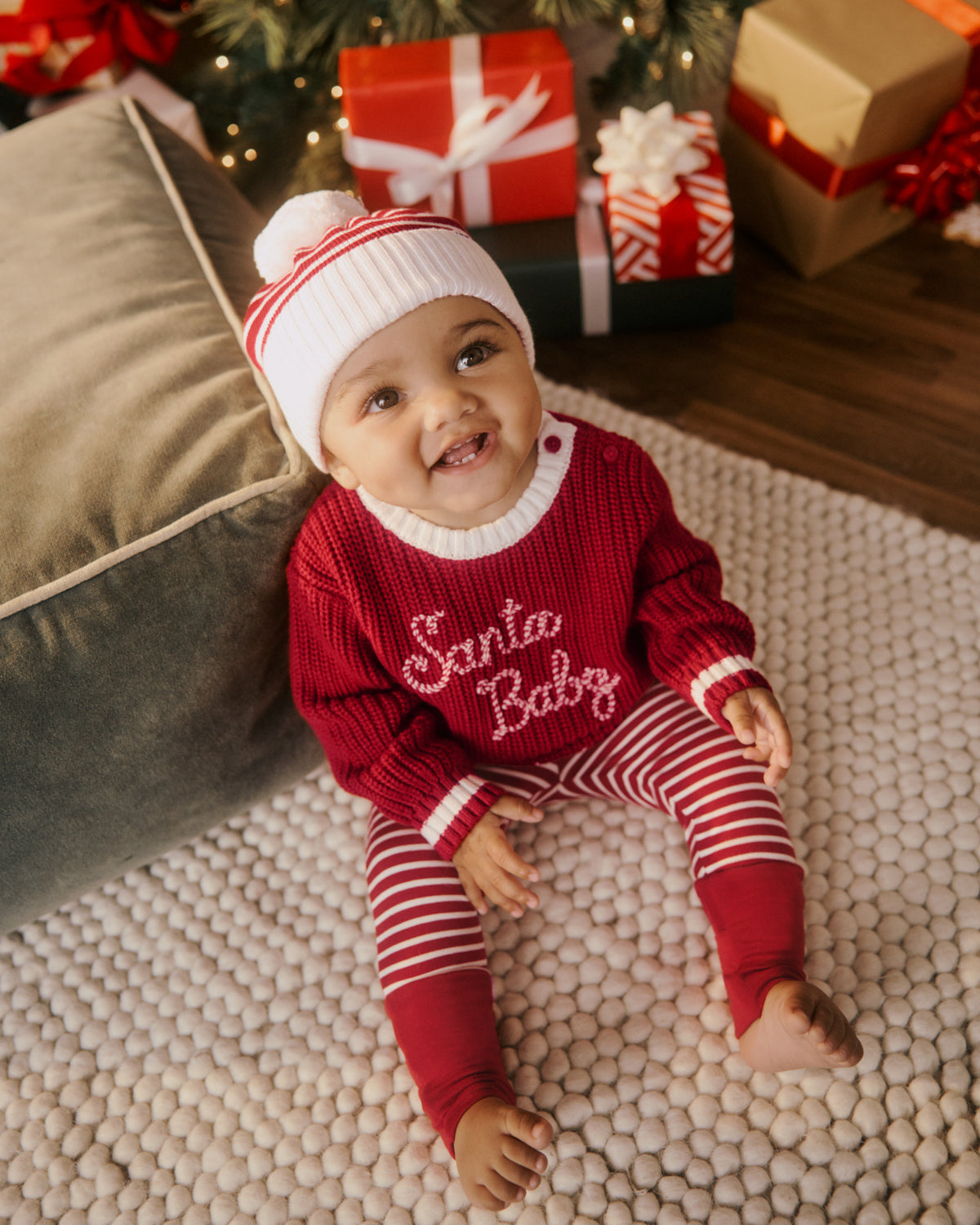 A baby wearing a red and white Christmas-themed knit sweater and striped leggings with presents and a Christmas tree in the background.