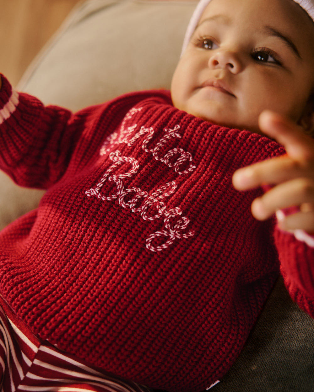 A close-up lifestyle shot of a baby wearing a red knit sweater with 'Santa Baby' written across the chest in white and red cursive embroidery.