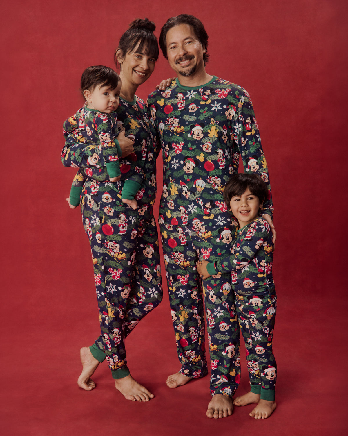 A family of four wearing matching dark green holiday pajamas with a colorful Mickey Mouse and friends print, standing against a solid red studio background.