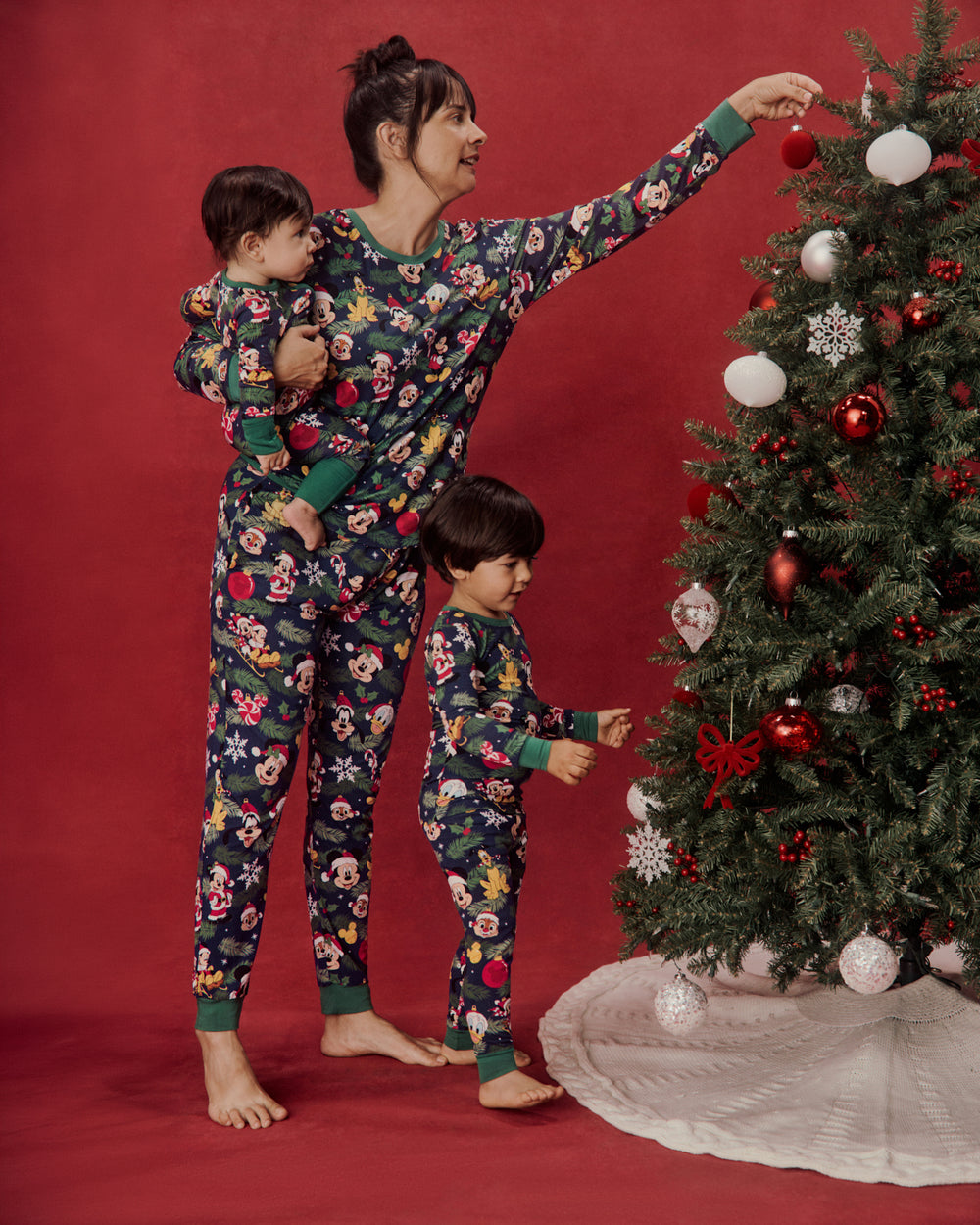 A woman and two children wearing matching floral Christmas pajamas pose next to a decorated Christmas tree against a solid red studio backdrop.
