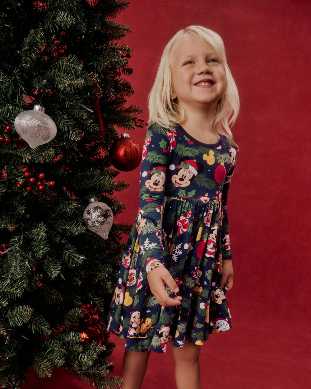 A young girl wearing a long-sleeve navy blue dress with a colorful Mickey Mouse and Minnie Mouse holiday print, standing next to a decorated Christmas tree against a solid red background.