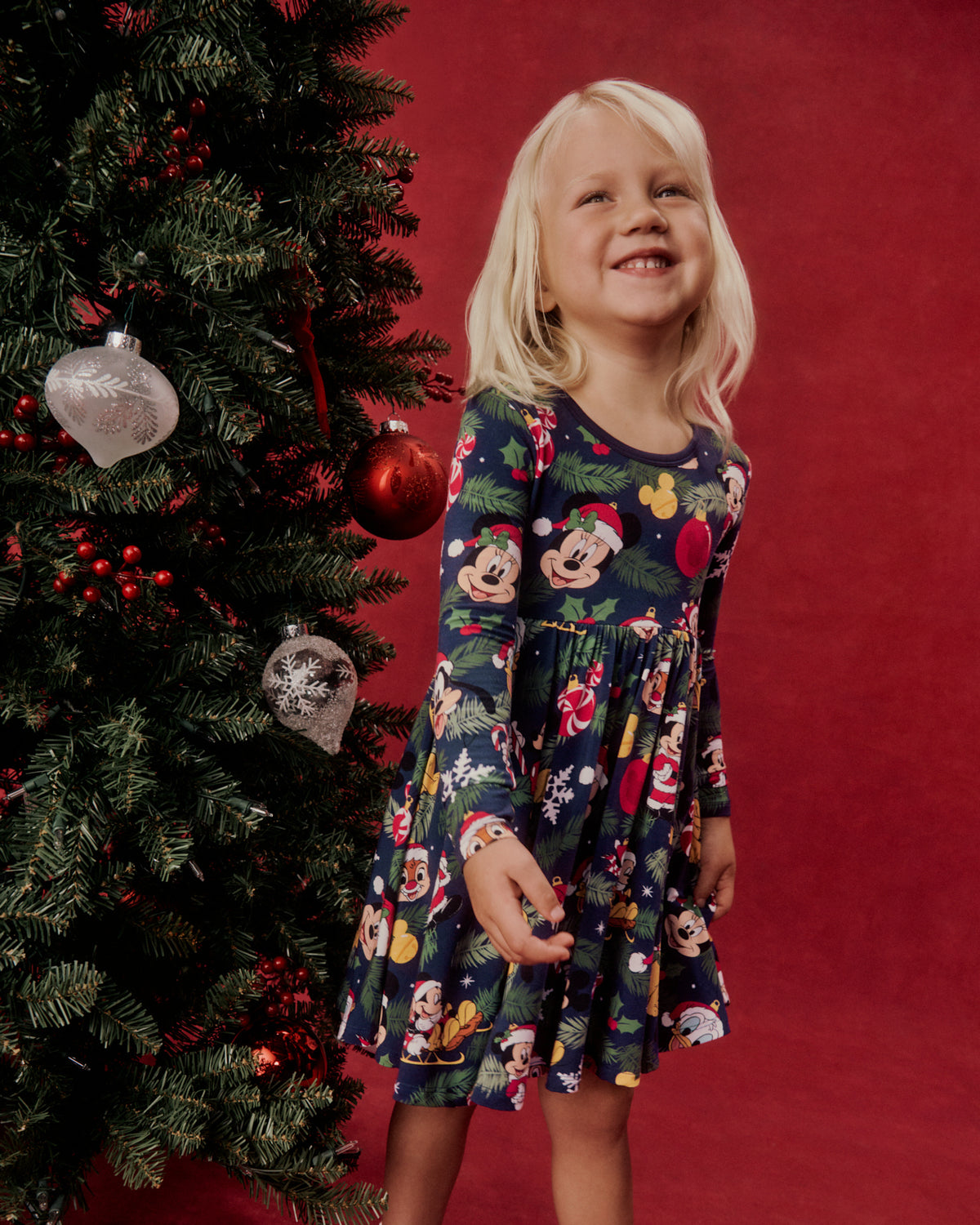 A young girl wearing a long-sleeve navy blue dress with a colorful Mickey Mouse and Minnie Mouse holiday print, standing next to a decorated Christmas tree against a solid red background.
