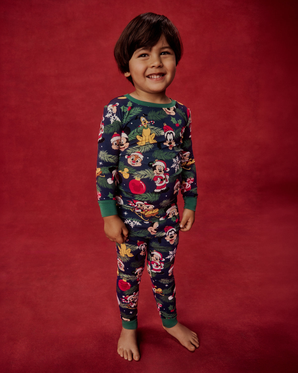 A young boy smiling and standing barefoot against a solid dark red studio backdrop, wearing a long-sleeve pajama set with a colorful Disney character print on a dark navy base.