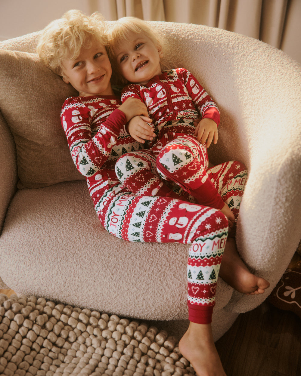 Two children sitting together on a beige armchair wearing matching red and white Christmas-themed long-sleeve pajamas with festive patterns and text.