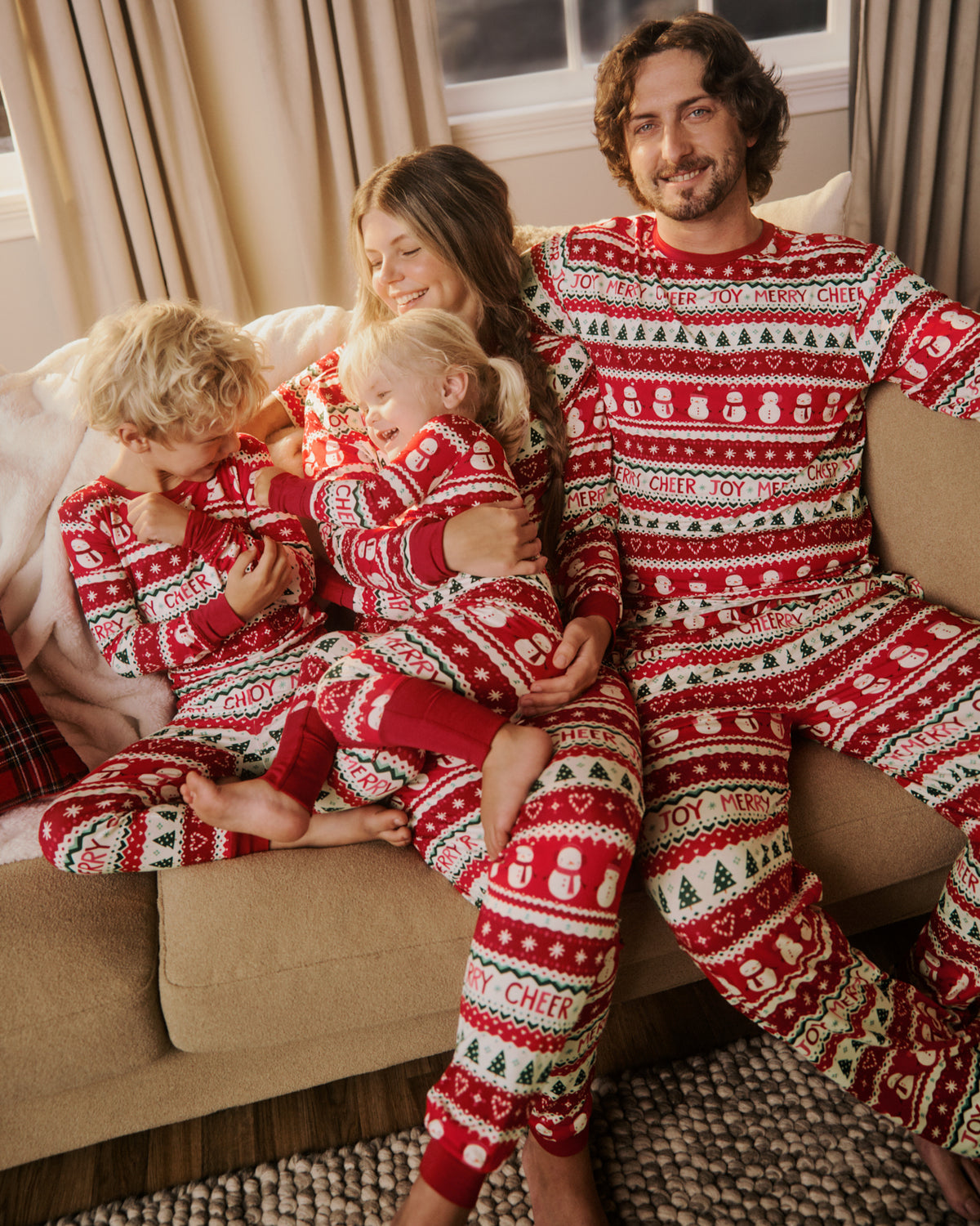A family of four wearing matching red and white Christmas-themed long-sleeve pajamas sits together on a beige couch in a cozy living room setting.