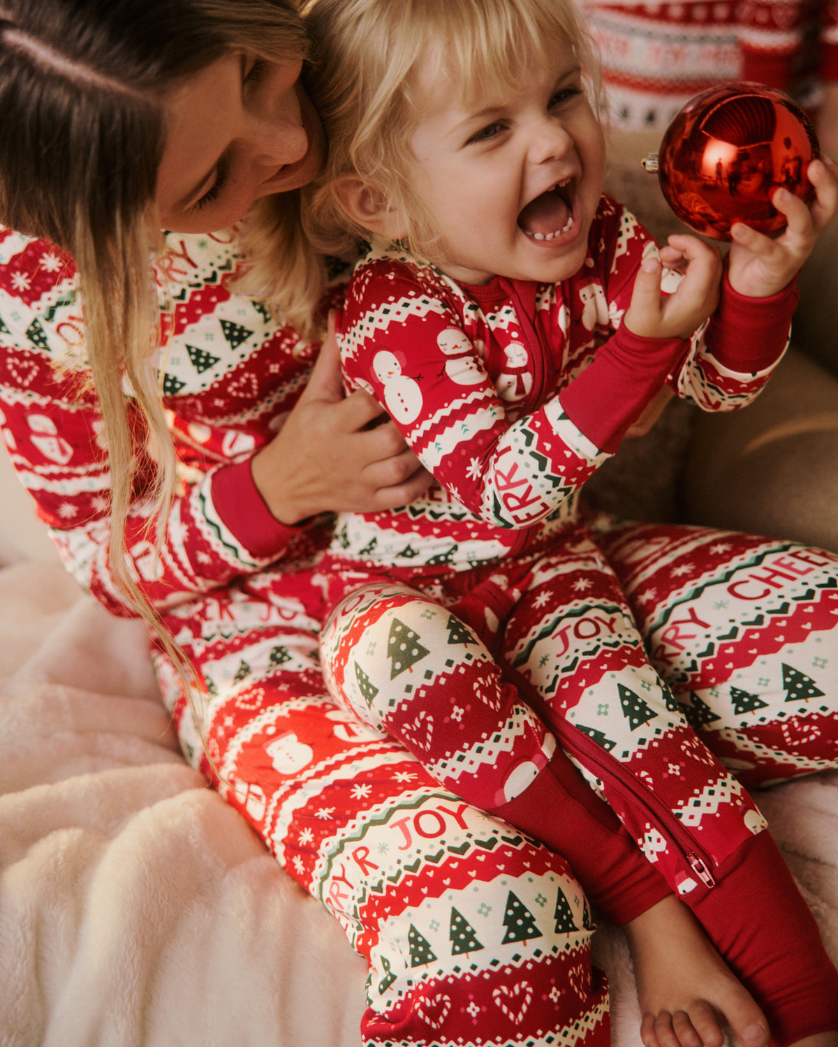 A woman and a young child wearing matching red and white Christmas-themed pajamas, sitting together on a soft blanket and holding a shiny red ornament.