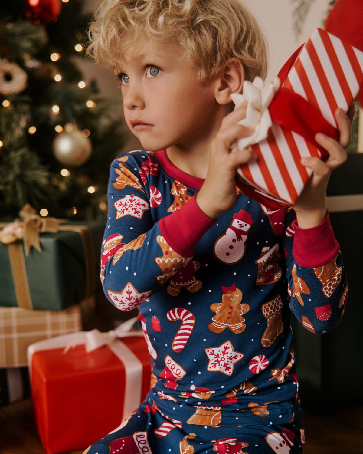 A young child wearing a festive blue Christmas pajama set with gingerbread and candy cane prints, holding a red and white striped gift box in front of a decorated Christmas tree.