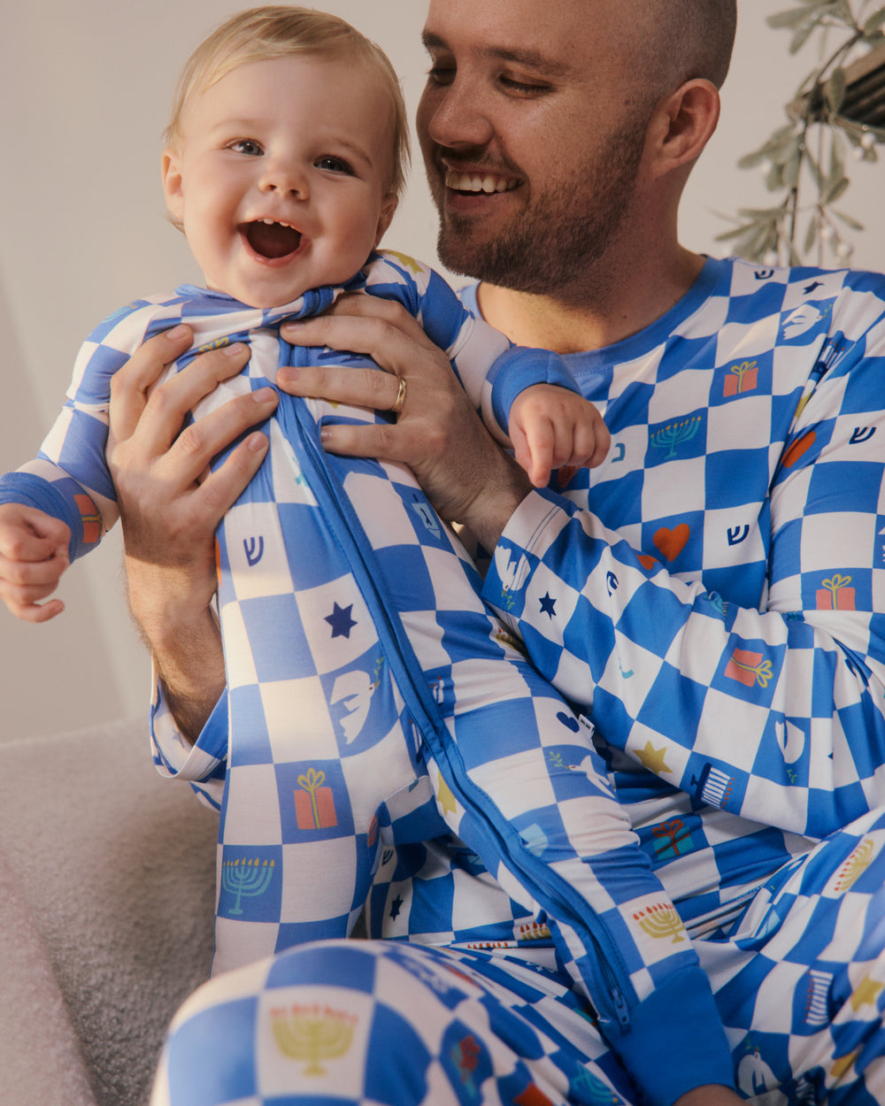 A smiling man and a baby wearing matching blue and white checkered pajamas with small colorful holiday and cultural motifs, sitting together indoors.