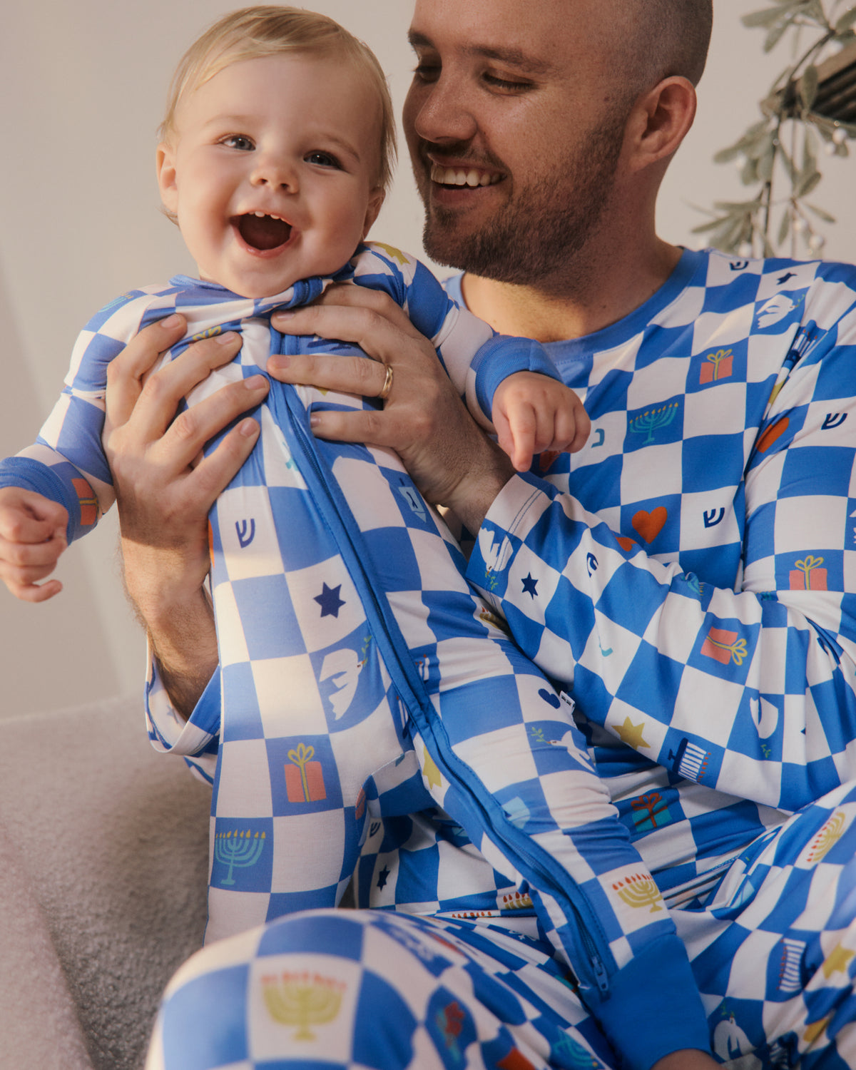 A smiling man and a baby wearing matching blue and white checkered pajamas with small colorful holiday and cultural motifs, sitting together indoors.