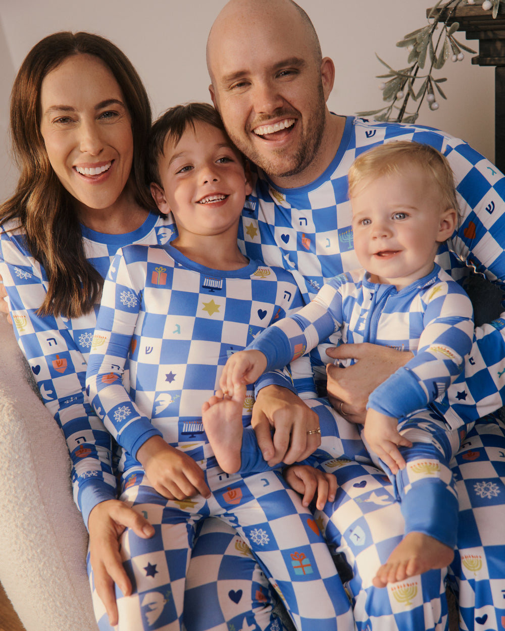 A family of four wearing matching blue and white checkered pajamas with Hanukkah icons, sitting together on a couch in a cozy indoor setting.