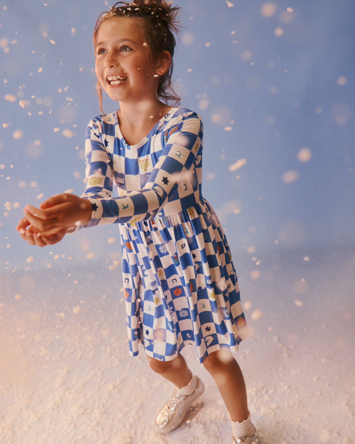 A young girl smiling and twirling in a blue and white checkered dress with Hanukkah icons, posed against a soft blue studio background with white snow-like particles.