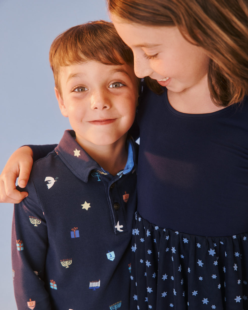 A young boy wearing a navy long-sleeve polo shirt with a colorful menorah and star pattern, standing next to a girl in a navy dress against a solid light blue background.