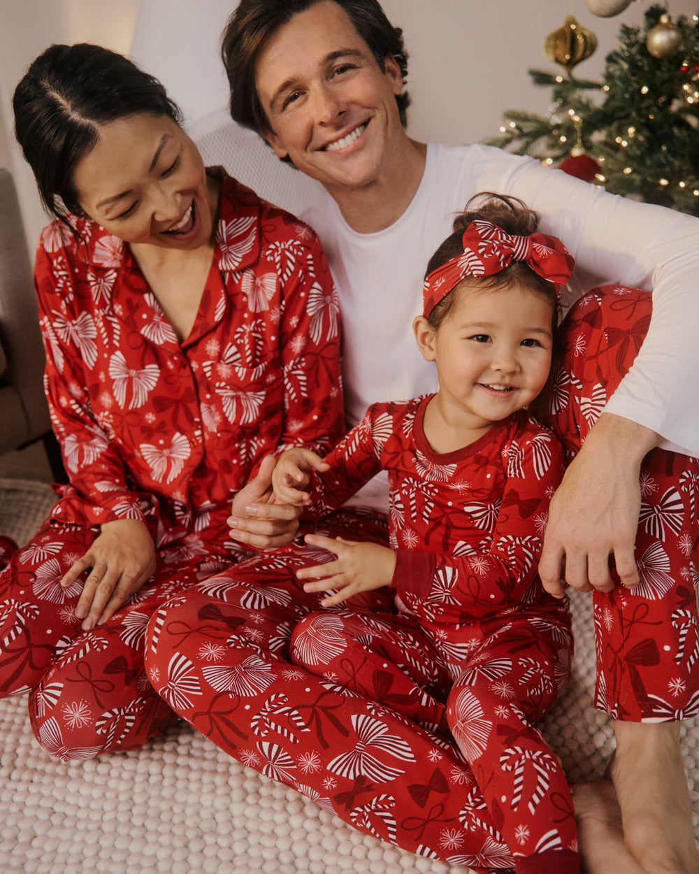 A family of three wearing matching red Christmas pajamas with white holiday ribbon patterns, sitting together on a cozy knit blanket in front of a decorated Christmas tree.