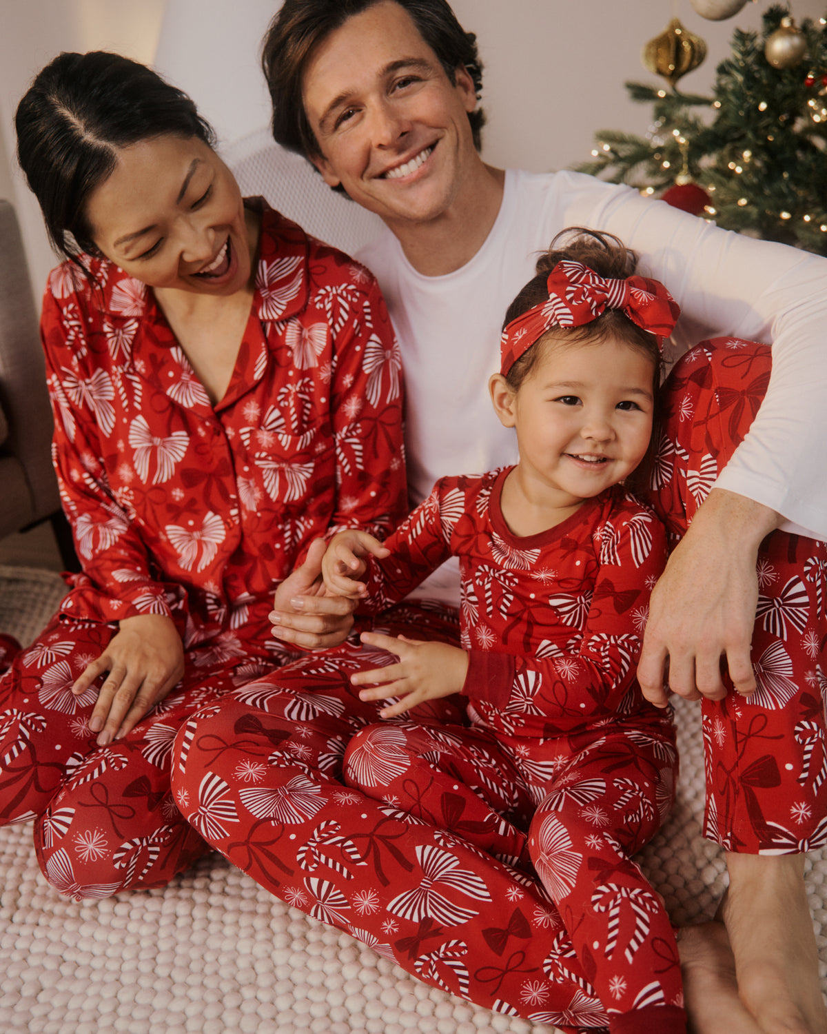 A family of three wearing matching red Christmas pajamas with white holiday ribbon patterns, sitting together on a cozy knit blanket in front of a decorated Christmas tree.