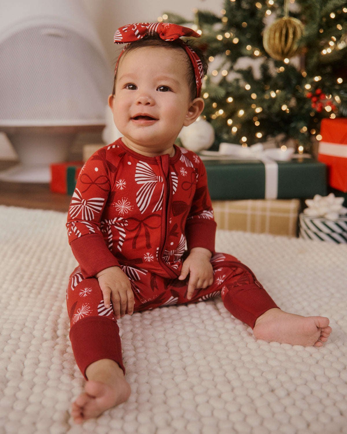 A baby wearing a red and white holiday print one-piece pajama set and a matching red bow headband, sitting on a white textured blanket in front of a decorated Christmas tree and wrapped gifts.