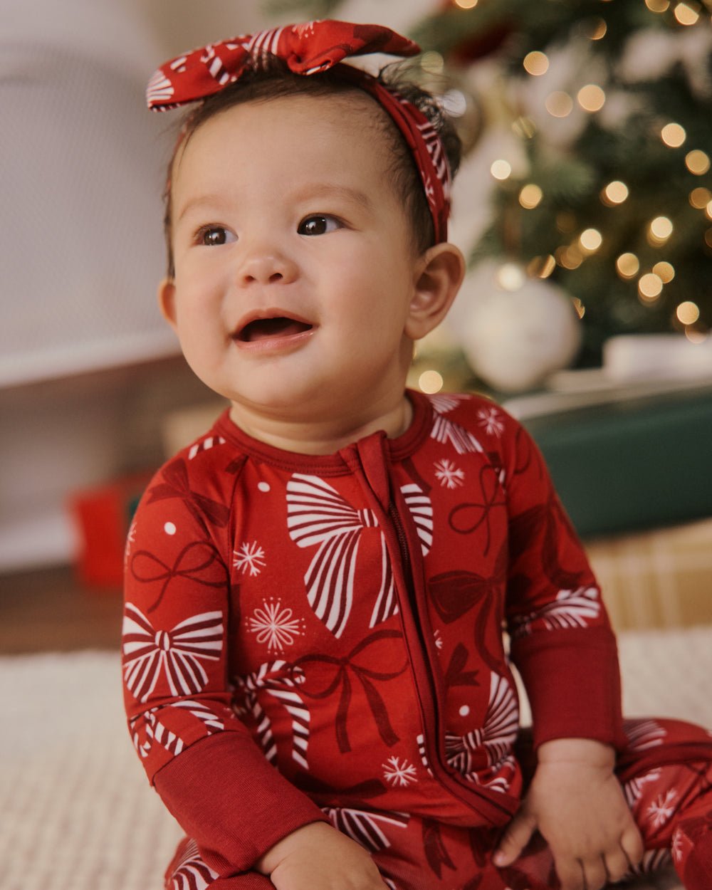 A baby wearing a festive red and white holiday print one-piece pajama and a matching red bow headband, sitting in front of a softly blurred Christmas tree with warm lights.